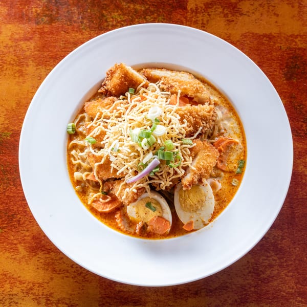 A white bowl filled with a rich orange curry, noodles, fried chicken, sliced eggs, and vegetables.