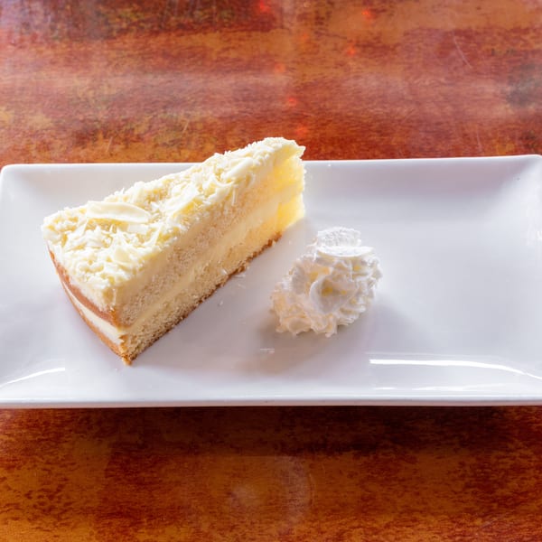 Slice of white chocolate cake with whipped cream on a white rectangular plate.