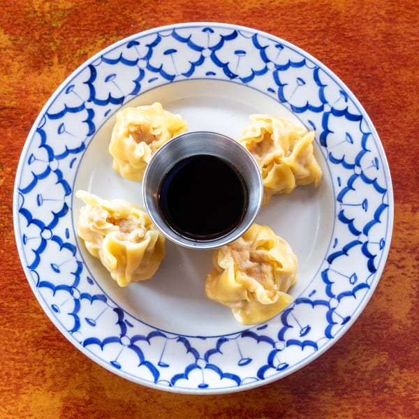 Four steamed dumplings with a small cup of soy sauce on a white and blue patterned plate.